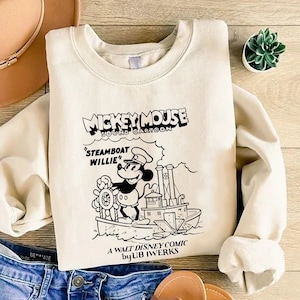 K&ouml;nnte beinhalten: Ein cremefarbener Pullover mit einer Schwarzwei&szlig;illustration von Mickey Mouse auf einem Dampfschiff. Der Text "Mickey Mouse Sound Cartoon" befindet sich &uuml;ber dem Bild und "Steamboat Willie" darunter. Der Text "A Walt Disney Comic by Ub Iwerks" befindet sich am unteren Rand des Bildes.