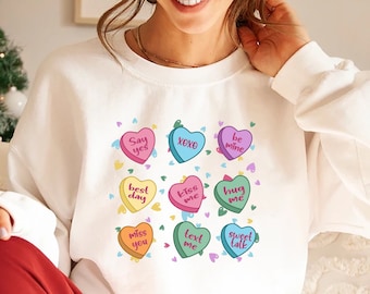 May include: White sweatshirt with a Valentine's Day theme featuring colorful hearts with phrases like "Say yes", "XOXO", "Best day", "Kiss me", "Hug me", "Miss you", "Text me", and "Sweet talk".