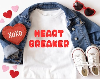 May include: White t-shirt with red text that says "HEART BREAKER" and a red felt heart with the text "XOXO" on it. The t-shirt is laying on a white surface with a denim jacket, white sneakers, and pink and red hearts.