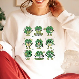 May include: White sweatshirt with a graphic of nine green four-leaf clovers reading books, drinking coffee, and holding flowers. Each clover has a smiling face.