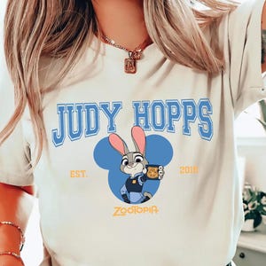 Op de afbeelding: Cr&egrave;mekleurig T-shirt met de tekst "JUDY HOPPS" in blauwe blokletters boven een cartoonafbeelding van Judy Hopps uit Zootopia. Het ontwerp bevat een blauw Mickey Mouse-silhouet en de tekst "EST. 2016" en "Zootopia".
