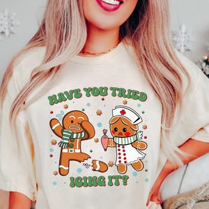 Könnte beinhalten: Cremefarbenes T-Shirt mit einem humorvollen Lebkuchen-Design. Die Grafik zeigt zwei Lebkuchenfiguren, eine mit gebrochenem Bein, und den Text "HAVE YOU TRIED ICING IT?". Das Design ist in Braun-, Grün- und Rottönen gehalten, ideal für die Weihnachtszeit.