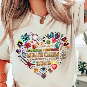 Op de afbeelding: Crèmekleurig T-shirt met een hartvormige afbeelding. De afbeelding bevat de tekst "Descendants Zombies + Camp Rock Worlds Collide Concert Tour 2026" en muziekinstrumenten, bloemen en andere kleurrijke elementen.