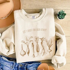 May include: Cream-colored sweatshirt with the phrase "Let's Root for Each Other" and a sepia-toned illustration of various root vegetables. The sweatshirt is paired with blue denim jeans, a tan hat, and sandals.