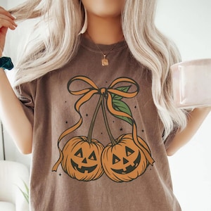 May include: Brown t-shirt featuring a pumpkin design. The graphic shows two orange jack-o'-lanterns with smiling faces, connected by a stem and bow. The shirt is a relaxed fit, perfect for Halloween or fall.