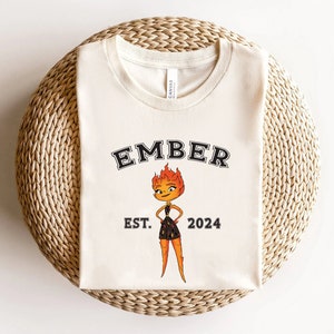 May include: A white t-shirt with a black graphic of a cartoon character with fire hair. The text "EMBER EST. 2024" is printed on the shirt.