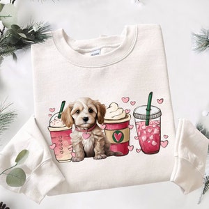 May include: A cream-colored sweatshirt with a graphic of a small, light brown dog sitting in front of three pink and white coffee cups with hearts and straws. The cups are decorated with hearts and the text "DOODOO".