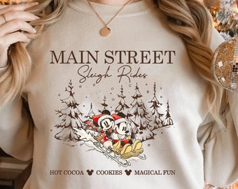 May include: A beige sweatshirt with a Christmas-themed design featuring Mickey Mouse and Minnie Mouse riding a sleigh in a snowy forest. The text "Main Street Sleigh Rides" is printed above the image, and "Hot Cocoa Cookies Magical Fun" is printed below.