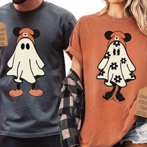 以下が含まれることがあります： 2枚のTシャツ。1枚はグレー、もう1枚はオレンジで、それぞれ漫画の幽霊のデザインが特徴です。幽霊はミッキーマウス風の帽子をかぶり、オレンジ色の靴を履いています。オレンジ色のシャツの幽霊には黒い花のディテールがあります。グレーのシャツには「COMFORT COLORS PEPPER COLOR」のタグが付いています。