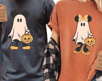 May include: A dark gray t-shirt featuring a ghost character with mouse ears holding a pumpkin bucket, and an orange t-shirt featuring a ghost character with mouse ears, a polka dot bow, and a pumpkin bucket. Both shirts have attached brand tags.