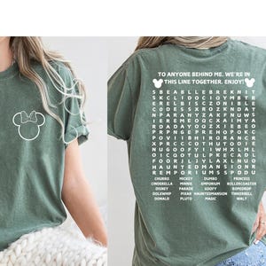 Puede incluir: Camiseta verde oliva con un contorno blanco de Minnie Mouse en la parte delantera. La parte trasera de la camiseta tiene el texto "TO ANYONE BEHIND ME, WE'RE IN THIS LINE TOGETHER. ENJOY!" y un juego de b&uacute;squeda de palabras de Disney.