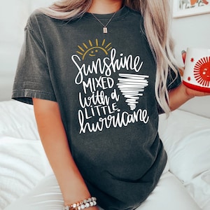 Op de afbeelding: Een donkergrijs T-shirt met een witte afbeelding waarop staat "Sunshine mixed with a little hurricane". De afbeelding bevat een gele zon en een witte tornado.