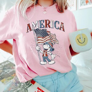 Peut inclure: T-shirt rose clair avec le mot "AMERICA" dans une police vieillie. Le t-shirt pr&eacute;sente un personnage de dessin anim&eacute; tenant un drapeau am&eacute;ricain. Le personnage porte une tenue patriotique. Le t-shirt est de couleur rose clair.