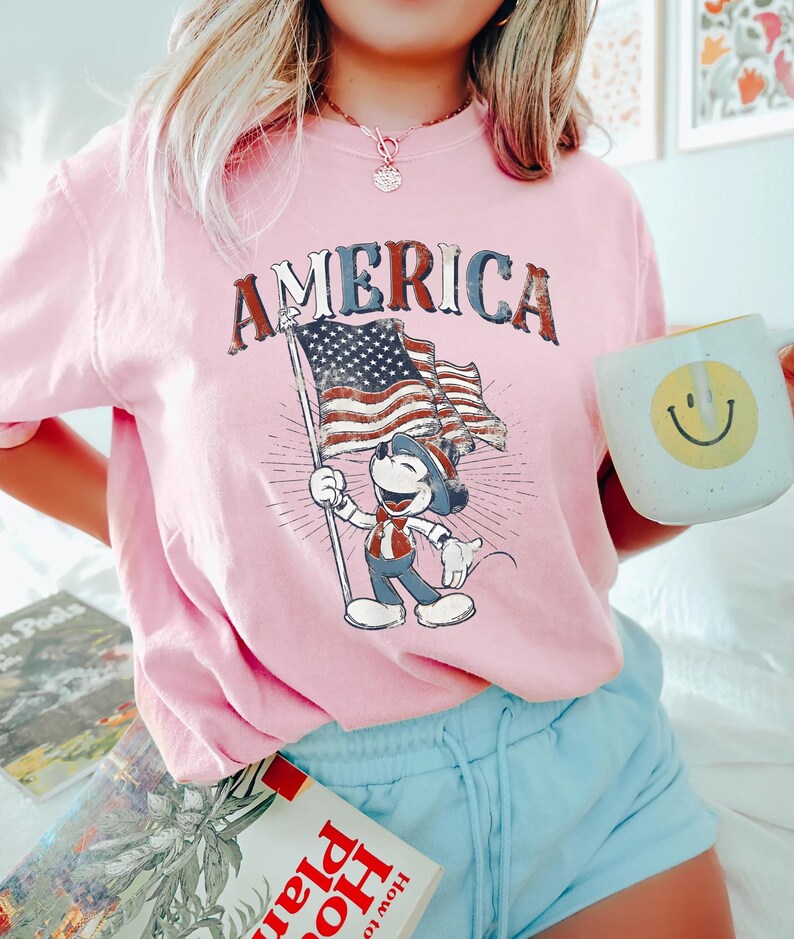 Peut inclure: T-shirt rose clair avec le mot "AMERICA" dans une police vieillie. Le t-shirt pr&eacute;sente un personnage de dessin anim&eacute; tenant un drapeau am&eacute;ricain. Le personnage porte une tenue patriotique. Le t-shirt est de couleur rose clair.