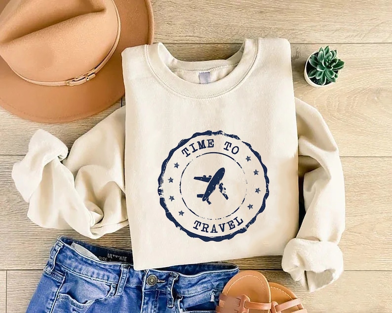 Puede incluir: Una sudadera de color crema con un sello azul desgastado que dice "Time to Travel" con un icono de avi&oacute;n dentro del sello.
