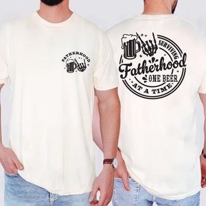 May include: Cream-colored t-shirt with a black graphic on the front and back. The front graphic says "Fatherhood" with a beer mug and skeleton hand. The back graphic says "Surviving Fatherhood One Beer At A Time."