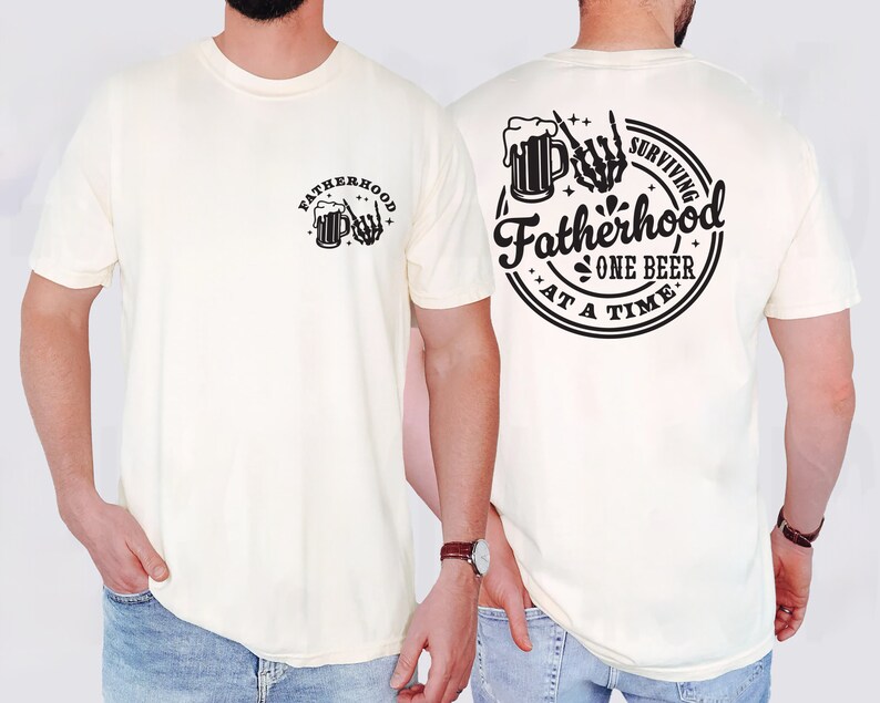 May include: Cream-colored t-shirt with a black graphic on the front and back. The front graphic says "Fatherhood" with a beer mug and skeleton hand. The back graphic says "Surviving Fatherhood One Beer At A Time."