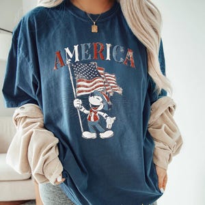 Peut inclure: T-shirt bleu marine avec le mot "AMERICA" en lettres rouges d&eacute;lav&eacute;es. Le t-shirt pr&eacute;sente un personnage de dessin anim&eacute; tenant un drapeau am&eacute;ricain. Le design a une esth&eacute;tique vintage et patriotique.