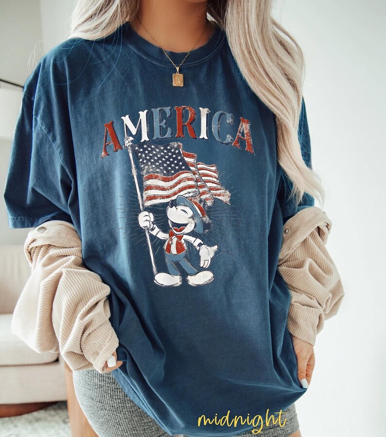 Peut inclure: T-shirt bleu marine avec le mot "AMERICA" en lettres rouges d&eacute;lav&eacute;es. Le t-shirt pr&eacute;sente un personnage de dessin anim&eacute; tenant un drapeau am&eacute;ricain. Le design a une esth&eacute;tique vintage et patriotique.