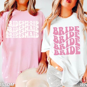 May include: Two t-shirts, one pink and one white, with a retro font design. The pink shirt says "BRIDESMAID" three times and the white shirt says "BRIDE" four times.