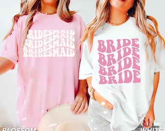Puede incluir: Dos camisetas, una rosa y una blanca, con un diseño de fuente retro. La camiseta rosa dice "BRIDESMAID" tres veces y la camiseta blanca dice "BRIDE" cuatro veces.