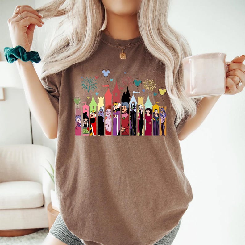 Puede incluir: Una camiseta marr&oacute;n con un gr&aacute;fico colorido de villanos de dibujos animados frente a un castillo. El gr&aacute;fico incluye fuegos artificiales y globos. La camiseta es de color neutro y tiene un ajuste relajado.