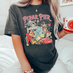 Puede incluir: Una camiseta negra con un gr&aacute;fico de estilo vintage de Pixar Pier en Disneyland. El gr&aacute;fico presenta personajes populares de Pixar como Buzz Lightyear, Woody, Mike Wazowski y Rayo McQueen. El texto "Pixar Pier" y "Disneyland" est&aacute;n incluidos en el gr&aacute;fico.