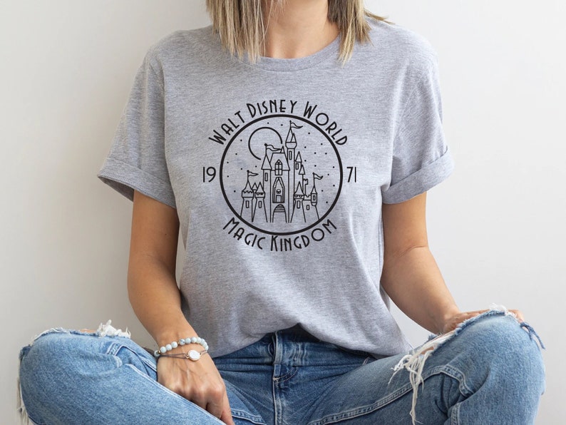 Puede incluir: Una camiseta gris con un gr&aacute;fico en blanco y negro del castillo de Cenicienta. El gr&aacute;fico incluye el texto "Walt Disney World" y "Magic Kingdom" con los a&ntilde;os "1971" a cada lado del castillo.