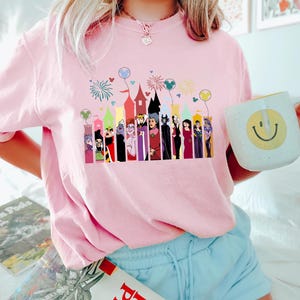 Puede incluir: Camiseta rosa claro con un dise&ntilde;o colorido de villanos de Disney y siluetas de castillos. El dise&ntilde;o incluye fuegos artificiales y globos de Mickey Mouse.