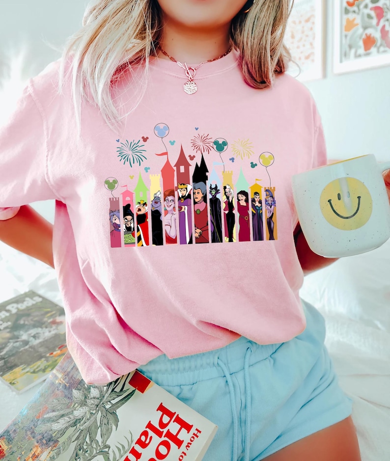 Puede incluir: Camiseta rosa claro con un dise&ntilde;o colorido de villanos de Disney y siluetas de castillos. El dise&ntilde;o incluye fuegos artificiales y globos de Mickey Mouse.