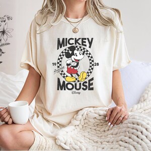 K&ouml;nnte beinhalten: Ein cremefarbenes T-Shirt mit einem Vintage-Mickey-Mouse-Motiv. Das Motiv zeigt Mickey Mouse in einem schwarz-wei&szlig; karierten Kreis mit dem Text "Mickey Mouse" und "Disney" darunter.