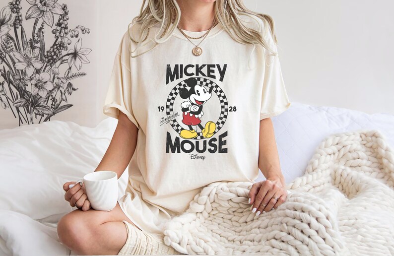 K&ouml;nnte beinhalten: Ein cremefarbenes T-Shirt mit einem Vintage-Mickey-Mouse-Motiv. Das Motiv zeigt Mickey Mouse in einem schwarz-wei&szlig; karierten Kreis mit dem Text "Mickey Mouse" und "Disney" darunter.