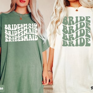 May include: Two t-shirts with a retro design. One is green with the word "BRIDESMAID" repeated in white. The other is ivory with the word "BRIDE" repeated in green.