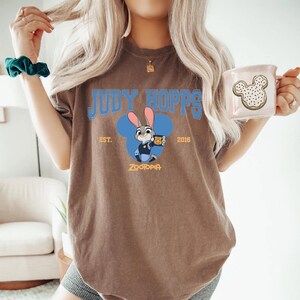 Op de afbeelding: Bruin t-shirt met een blauwe afbeelding van Judy Hopps uit Zootopia, met de tekst "JUDY HOPPS" boven het personage en "EST. 2016" aan weerszijden. Een witte mok met een Mickey Mouse koekjesontwerp wordt in de hand gehouden.