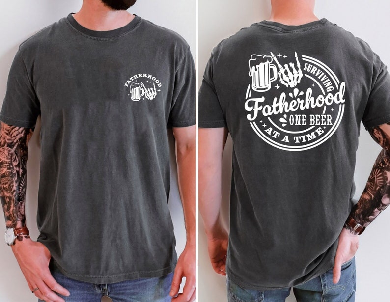May include: Dark gray t-shirt with white graphic text and illustrations. The front features the word "Fatherhood" with a beer mug and skeleton hand. The back says "Surviving Fatherhood One Beer At A Time" in a circular design with a beer mug and skeleton hand.