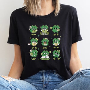 May include: A black t-shirt with a graphic of nine green four-leaf clovers with faces. Each clover is doing a different activity, such as reading a book, drinking coffee, or holding a flower.