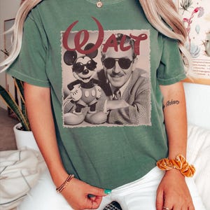 May include: A moss green short-sleeve t-shirt featuring a graphic print of Walt Disney and Mickey Mouse both wearing sunglasses. The text WALT is printed in a large, dark red font above the black and white image of the two figures.
