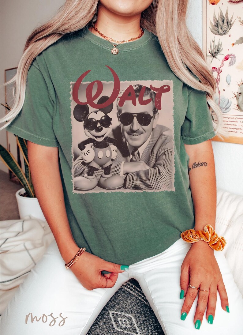 May include: A moss green short-sleeve t-shirt featuring a graphic print of Walt Disney and Mickey Mouse both wearing sunglasses. The text WALT is printed in a large, dark red font above the black and white image of the two figures.