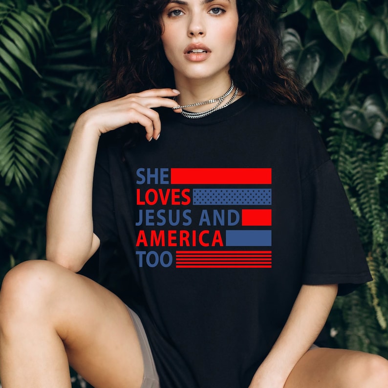 Może przedstawiać: Czarny T-shirt z czerwono-biało-niebieskim nadrukiem, na kt&oacute;rym widnieje napis "SHE LOVES JESUS AND AMERICA TOO".