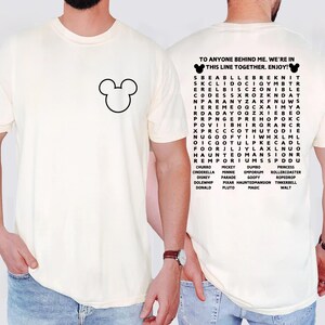 May include: Cream-colored t-shirt featuring a black Mickey Mouse outline on the front. The back of the shirt displays a word search puzzle with Disney-related words and the text "TO ANYONE BEHIND ME, WE'RE IN THIS LINE TOGETHER. ENJOY!"
