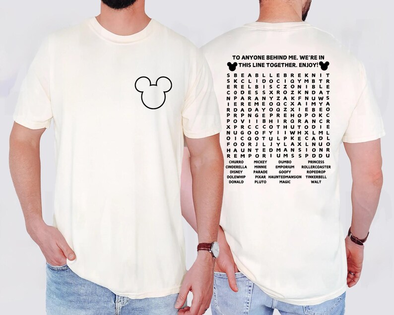 May include: Cream-colored t-shirt featuring a black Mickey Mouse outline on the front. The back of the shirt displays a word search puzzle with Disney-related words and the text "TO ANYONE BEHIND ME, WE'RE IN THIS LINE TOGETHER. ENJOY!"