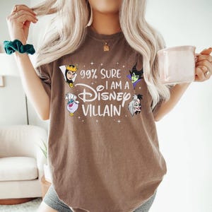 以下が含まれることがあります： 「99% Sure I am a Disney Villain」の文字とディズニーヴィランズのイラストがプリントされた茶色のTシャツ。カジュアルなスタイルで、普段使いに最適です。
