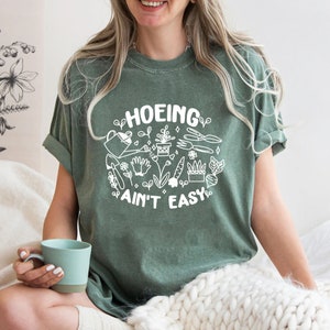 Op de afbeelding: Een groen T-shirt met een wit grafisch ontwerp met de tekst "HOEING AIN'T EASY" en afbeeldingen van tuingereedschap en planten.