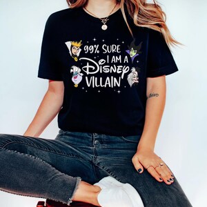 以下が含まれることがあります： 「99% Sure I am a Disney Villain」の文字とディズニーヴィランズのイラストがプリントされた黒いTシャツ。青いジーンズと白い靴を履いた人が赤いスツールに座っています。