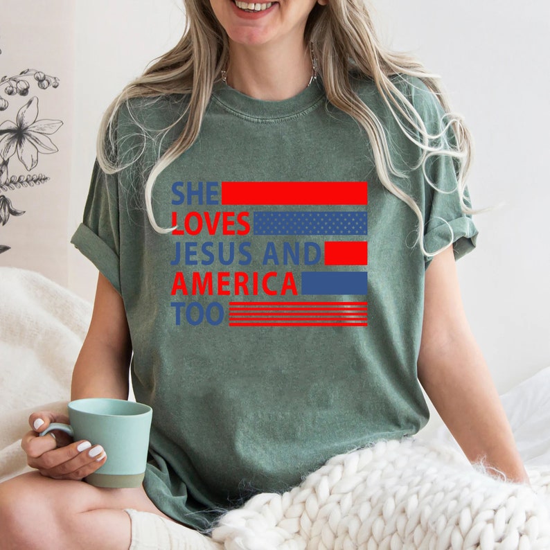 Może przedstawiać: Zielona koszulka z napisem "SHE LOVES JESUS AND AMERICA TOO" w czerwonych i niebieskich literach. Tekst jest ułożony pionowo z czerwonymi i niebieskimi paskami z tyłu.