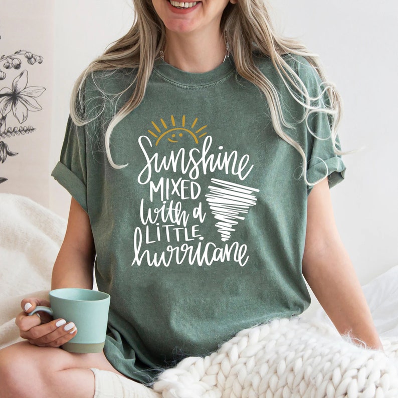 Op de afbeelding: Een groen T-shirt met een witte afbeelding waarop staat "Sunshine mixed with a little hurricane". De afbeelding bevat ook een gele zon en een witte tornado.
