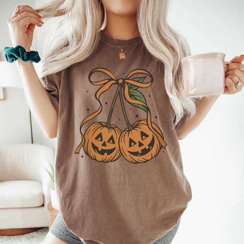 May include: Brown t-shirt featuring a pumpkin design. The graphic shows two orange jack-o'-lanterns with smiling faces, connected by a stem and bow. The shirt is a relaxed fit, perfect for Halloween or fall.