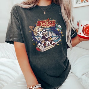 May include: A dark gray t-shirt with a vintage-style graphic of Mickey Mouse and Donald Duck riding a spaceship in space. The graphic includes the text "Space Mountain".