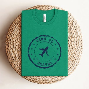 Puede incluir: Una camiseta verde con un dise&ntilde;o de sello azul que dice "Time to Travel" con un avi&oacute;n y una tabla de surf.