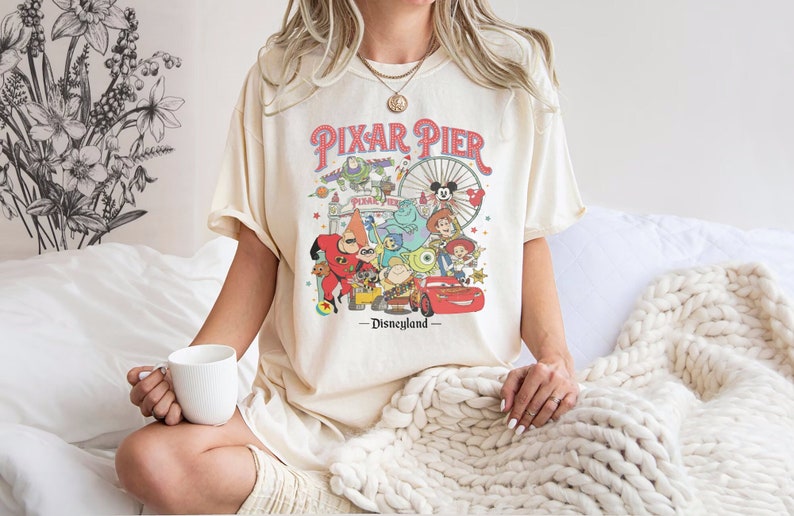 Puede incluir: Una camiseta blanca con un gr&aacute;fico de estilo vintage de Pixar Pier en Disneyland. El gr&aacute;fico presenta personajes de pel&iacute;culas populares de Pixar, incluyendo Buzz Lightyear, Woody, Mike Wazowski, Sulley y Mr. Incre&iacute;ble. El texto "Pixar Pier - Disneyland" est&aacute; impreso debajo del gr&aacute;fico.